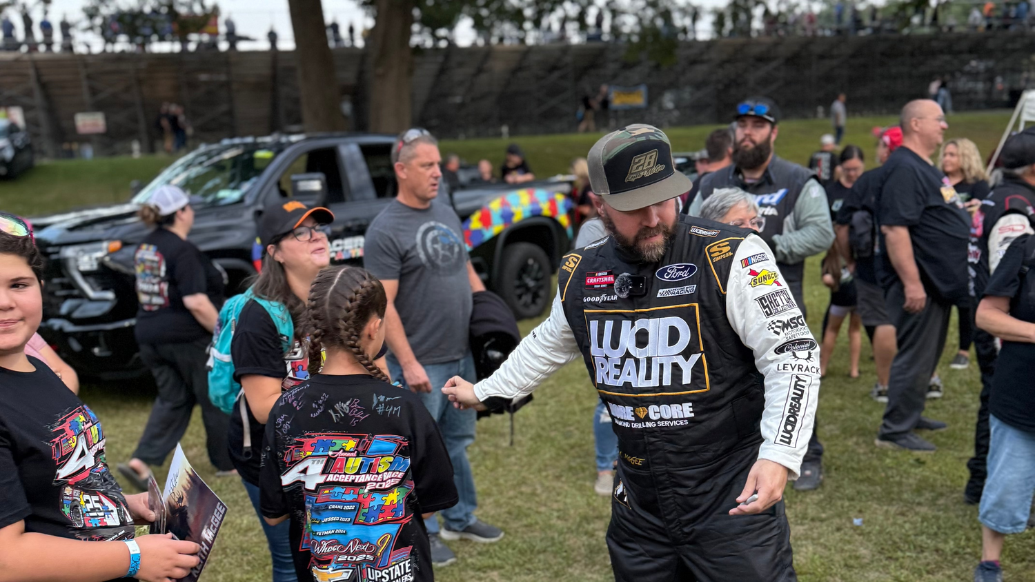 Man in racing gear shaking hands with a young fan at an outdoor event.
