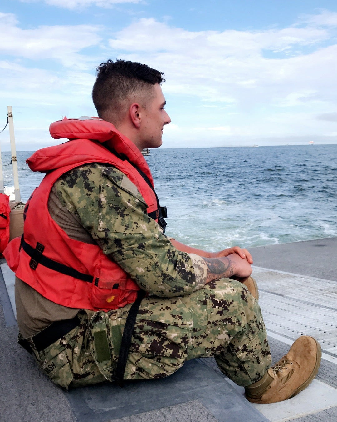 Person in military uniform and life vest sitting on a boat deck with ocean and sky in the background