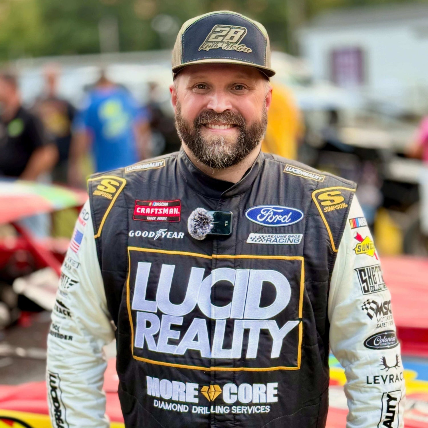 Man wearing a racing suit with various sponsor logos at a race track.